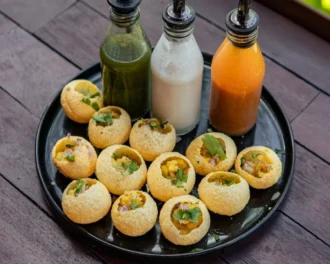 Puffed pani puri shells filled with potato and chickpea on a black plate, with three bottles of flavoured pani