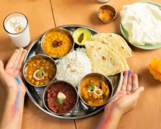 Steel thali of dal, chickpea curry, rice, halwa and roti with a lassi, framed by colour-stained hands