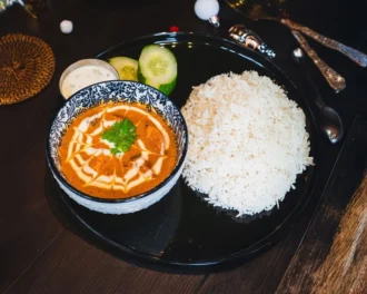 Butter chicken in a patterned bowl with steamed basmati rice, cucumber and lime on a black plate